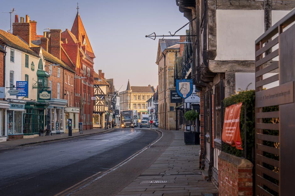 A charming English town centre street at dusk with warm lights glowing from shop windows and historic buildings