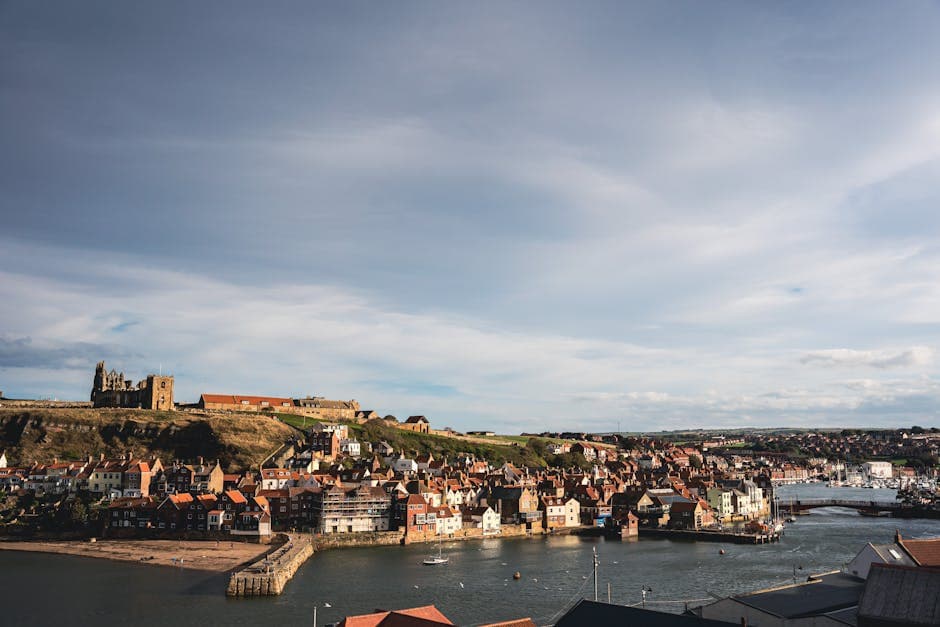 A picturesque coastal town with historic ruins on a hilltop, a river running through it, and a wide sky above