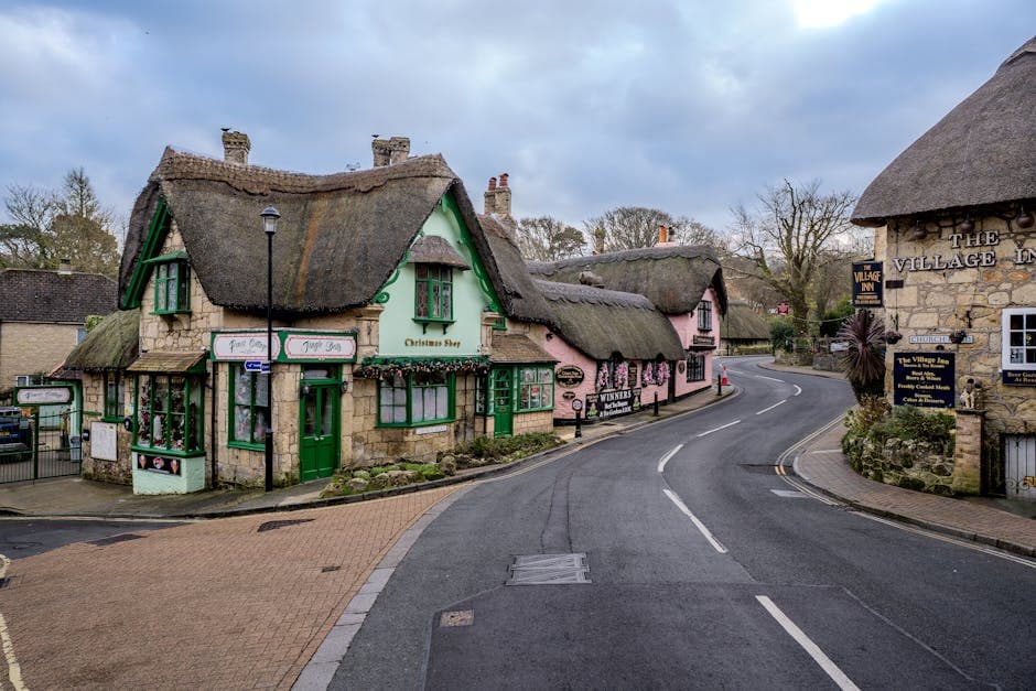 A charming English village high street with traditional thatched buildings, independent shops, and flower-lined pavements